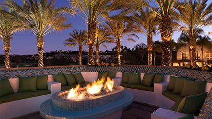 A beautiful outdoor patio area with a fire pit in the center. The fire pit is made of blue tiles and is surrounded by green cushioned sofas. The patio is decorated with palm trees and other tropical plants. In the background there is a view of a city skyline at night. The sky is dark and the overall atmosphere is peaceful and serene.