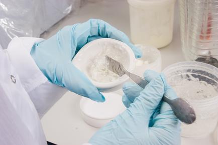 A person's hands wearing blue gloves holding a small white bowl with a spoon in it. The person is wearing a white lab coat and appears to be in a laboratory setting. The bowl is filled with a white substance possibly a mixture of flour or other ingredients and there are several other bowls and containers in the background. The background is blurred but it seems like the person is working in a scientific or laboratory setting with other laboratory equipment.