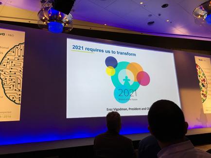 A large screen in a conference room with a presentation on it. The screen displays the text "2021 requires us to transform 2021" in bold colorful circles. Below the text there is an image of a circuit board with the year 2021 written on it indicating that the presentation is about the future of the company. On the right side of the screen there are two smaller images of the same circuit board one on the left side and one in the center. The room is dimly lit with blue and purple lights and there are three people sitting in front of the projector screen attentively listening to the presentation.
