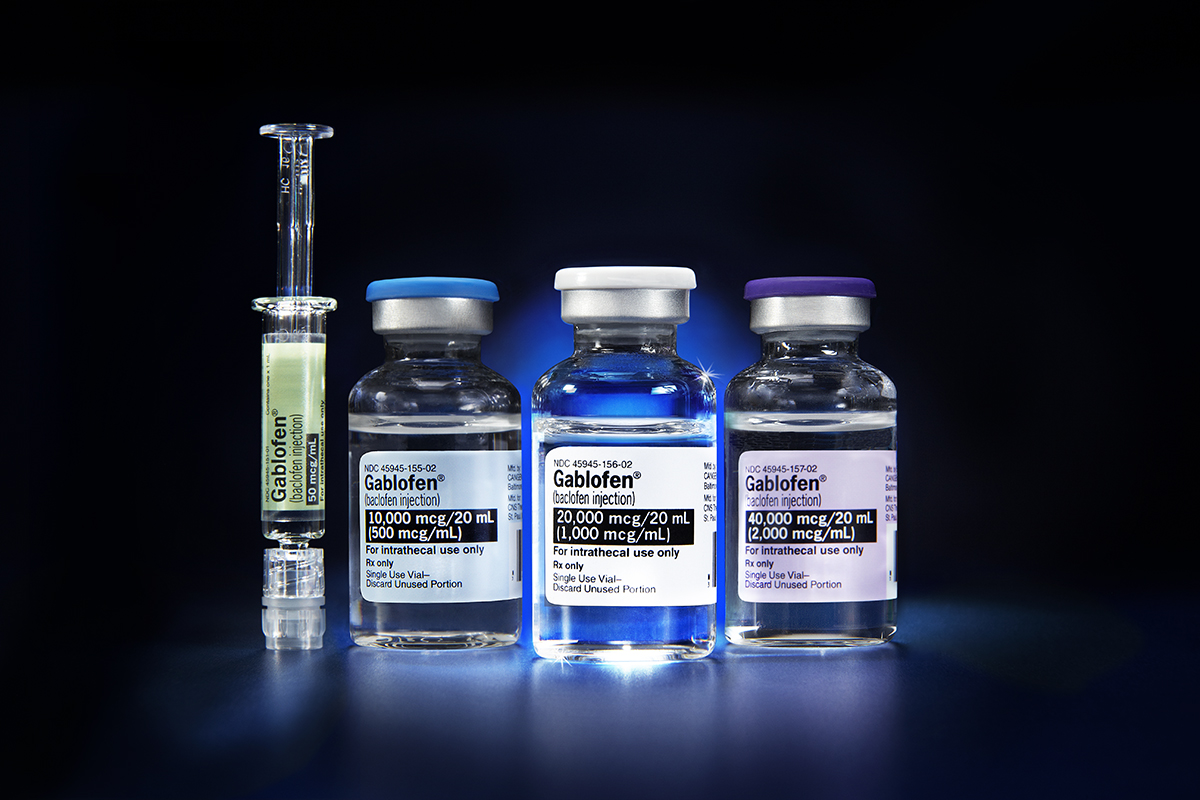 A prefilled syringe and three small glass vials of Gablofen (baclofen injection) in various dosages (50 mcg/mL 500 mcg/mL 1000 mcg/mL and 2000 mcg/mL from left to right). All are photographed against a dark background which is primarily black with a blue spotlight shining through the syringe and vials (from behind).