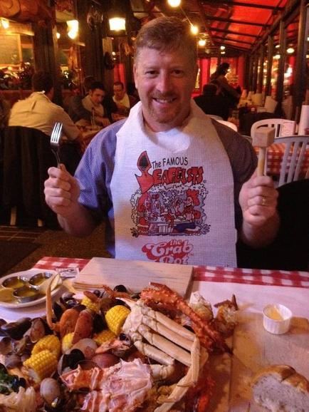 A man sitting at a table in a restaurant. He is wearing a white apron with the words "The Famous seafood" printed on it. He has a big smile on his face and is holding a fork in his right hand. On the table in front of him there is a large platter of seafood including lobster clams corn and mussels. There is also a small bowl of sauce on the table. The restaurant has a red and white checkered tablecloth and there are other tables and chairs in the background.