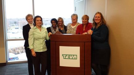 A group of nine women standing behind a wooden podium with the word "tea" written on it. They are all smiling and appear to be posing for a photo. The podium is in an office setting with a large window in the background through which we can see a view of a parking lot and trees. The women are of different ages and ethnicity's and they are all dressed in formal attire. The woman on the left is wearing a green blouse and black pants the woman in the center is wearing glasses and the woman next to her is wearing an orange blouse. All of them are looking at the camera and smiling.