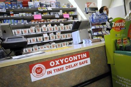 A pharmacy counter with a sign that reads "Oxycontin in time delay safe". The counter is made of wood and has a cash register and a printer on it. Behind the counter there are shelves stocked with various medicines and products. On the right side of the image there is a green sign with the logo of Zyrtec and the text "The Fastest 24-Hour Allergy Relief". A woman is standing behind the counter and appears to be paying for the medication.