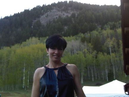 A young woman standing in front of a mountain with trees in the background. She is wearing a blue sleeveless dress with a halter neckline and has short black hair styled in a short bob. The woman is smiling and looking directly at the camera. In the background there is a white tent and a wooden structure. The sky is overcast and the overall mood of the image is peaceful and serene.