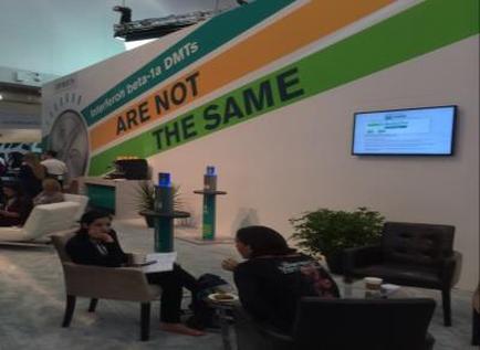A trade show booth with a large banner that reads "Are not the same". The booth is decorated with a green and orange color scheme and has a large screen on the right side. There are two people sitting in chairs in front of the booth engaged in conversation. On the left side there is a desk with a computer and other items on it. The booth appears to be well-lit with natural light coming in from the ceiling.