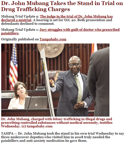 Dr. John Mubang a judge in the trial of Dr. Johns ubangi standing in front of a podium with an American flag in the background. He is wearing a suit and tie and is gesturing with his right hand as he speaks. Behind him there is a woman sitting at a desk with a laptop and a microphone. The image is accompanied by text that reads "Dr. John bang takes the Stand in Trial on Drug Trafficking Charges".