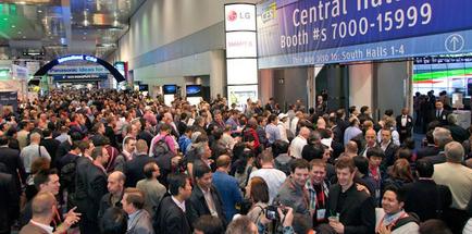 A large crowd of people gathered in a convention hall. The hall is filled with people of different ages genders and ethnicity's. The people are walking around and interacting with each other. On the right side of the image there is a large blue sign that reads "Central Hall Booth #s 7000-15999". In the background there are several other signs and banners hanging from the ceiling. The overall atmosphere appears to be lively and bustling.