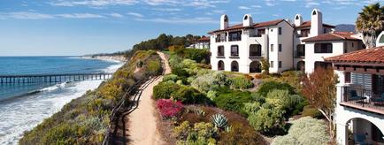 A beautiful view of a coastal area with a row of white buildings on the right side. The buildings have arched windows and balconies and are surrounded by lush greenery and colorful flowers. On the left side of the image there is a sandy beach with a pier extending into the ocean. The sky is blue and the water is a deep blue-green color. In the background there are mountains and a clear blue sky. The overall scene is peaceful and serene.