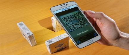 A person's hand holding a Samsung smartphone with a QR code on the screen. The QR code is displayed on the phone's home screen and there are three small white boxes with the same QR code printed on them. The boxes are placed on a wooden surface. The background is blurred but it appears to be a wooden table. The person is likely scanning the QR code for a product or service.