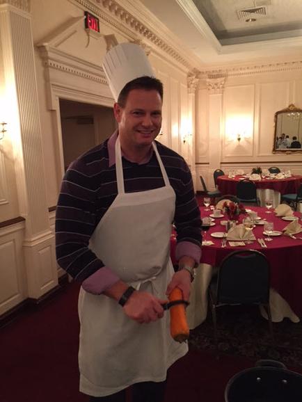 A man wearing a white chef's hat and a white apron. He is standing in a large room with a high ceiling and ornate decorations. The man is smiling and holding a carrot in his right hand. He appears to be in the middle of a meal as there are tables and chairs set up in the background with red tablecloths and white tablecloth. There is a mirror on the wall and a chandelier hanging from the ceiling. The overall atmosphere of the room is elegant and sophisticated.