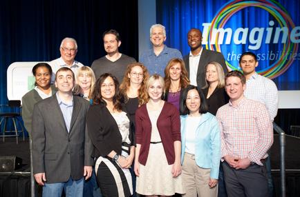 A group of people standing on a stage with a blue curtain in the background. There is a large screen behind them with the words "Imagine the possibilities" written in colorful letters. The people in the group are of different ages genders and ethnicity's. They are all smiling and appear to be posing for a group photo. Some of them are wearing casual clothing while others are dressed in formal attire. The overall mood of the image is happy and confident.