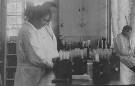 A black and white photograph of two women in a laboratory. They are both wearing lab coats and appear to be working on a project. The woman on the left is standing in front of a table with several test tubes and other scientific equipment on it. She is holding a small object in her hands and appears to be examining it closely. The man on the right is sitting at the table and is looking at the test tubes. There is a window in the background with curtains and a shelf with various scientific equipment. The photograph appears to have been taken in the early 20th century.