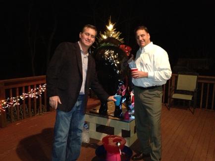 Two men standing on a wooden deck at night. They are both smiling and appear to be posing for the photo. The man on the left is wearing a black blazer and jeans while the man in the white shirt is holding a red cup. In the background there is a large Christmas tree decorated with lights and ornaments. On the right side of the image there are two chairs and a table with various items on it. The deck is lit up with string lights creating a warm and cozy atmosphere.