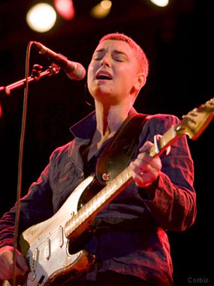 A young man playing an electric guitar on stage. He is wearing a dark blue shirt and appears to be singing into a microphone. The background is dark and there are red and yellow lights shining down on him. The man is holding the guitar with both hands and his eyes are closed as he plays. He seems to be in the middle of a performance.