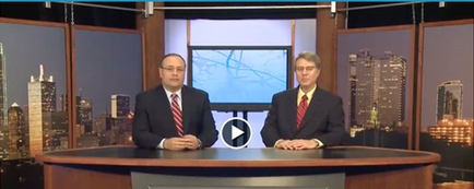 Two men sitting at a desk in a news studio. They are both wearing suits and appear to be in the middle of a news broadcast. The desk is in front of a large window with a view of a city skyline in the background. The man on the left is bald and wearing glasses while the man on on the right is wearing a suit and tie. Both men are looking directly at the camera with serious expressions on their faces. There is a play button in the center of the image indicating that they are in the midst of a conversation.