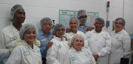A group of nine people nine women and one man standing in a laboratory. They are all wearing white lab coats with green labels on them and hairnets on their heads. The woman in the center is wearing a blue shirt and is smiling at the camera. Behind them there is a whiteboard with the words "PLAN DO NOT ENTER" written on it. The people in the group appear to be of different ages and ethnicity's. There is a computer monitor and other equipment in the background.