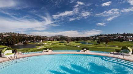 A beautiful landscape of a golf course with a large swimming pool in the foreground. The pool is rectangular in shape and has a blue-green color. There are two lounge chairs on either side of the pool with green cushions providing a perfect spot to relax and enjoy the view. In the background there are several golf courses with lush green grass and trees. The sky is blue with white clouds scattered across it and there are a few buildings and houses visible in the distance. The overall atmosphere of the image is peaceful and serene.