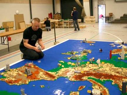 A man kneeling on the floor of a large room with a large map spread out in front of him. The map appears to be made up of various Lego pieces including buildings trees and other structures. The man is wearing a black t-shirt and is holding a small object in his hand. In the background there are other people working on the map and there are tables and benches scattered around the room. The room has a high ceiling with large windows and a door on the right side.