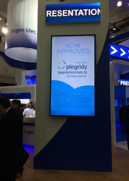 A large blue and white signboard with the word "presentation" written in bold letters at the top. Below the signboard there is a large screen with the text "Now Approved" and "pleurisy" in white letters. The signboard is located in an exhibition hall with other booths and people in the background. The overall theme of the image is clean and modern.