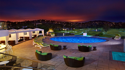 A beautiful outdoor swimming pool area at night. The pool is rectangular in shape and has a blue-green color. There are several lounge chairs and sofas arranged around the pool providing a comfortable seating area for people to relax and enjoy the view. On the left side of the image there is a white gazebo with a curved roof providing shade for the pool area. In the background there are trees and hills and the sky is dark indicating that it is either dusk or early morning. The overall atmosphere of the space is peaceful and relaxing.