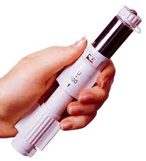 A hand holding a white medical device with a black-colored section possibly a chamber. The device has a cylindrical shape with a pointed end and a small opening at the top. It appears to be related to the dermal administration of medications. The body of the device is made of plastic and has a textured surface. The background is plain white.