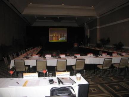 A large conference room with a long table set up for a meeting. The table is covered with a white tablecloth and has nameplates glasses and other items on it. There are several chairs around the table and a projector screen in the background. The room has a high ceiling with recessed lighting and a carpeted floor. The walls are painted in a dark color and there are plants on either side of the room.