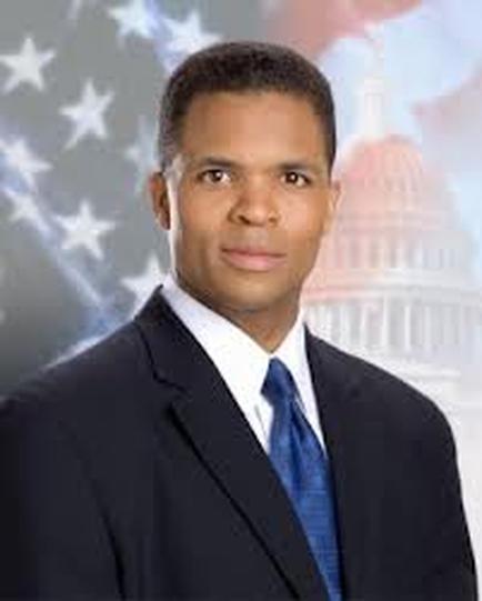 A professional headset of a man in a suit and tie. He is standing in front of an American flag and the Capitol building in the background. The man is African-American and appears to be in his late 30s or early 40s. He has short dark hair and is looking directly at the camera with a serious expression on his face. The image appears to have been taken in a professional setting.