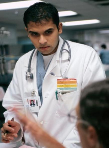 A young man in a white lab coat with a stethoscope around his neck. He is sitting at a desk and appears to be engaged in a conversation with another person. The man is holding a pen and seems to be explaining something to the other person who is sitting in front of him. The background is blurred but it seems like the focus is on the two people in the foreground. The image appears to have been taken in a hospital or medical facility.