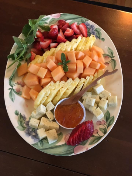 Of a platter of fruit and cheese. The platter is white with a green floral design around the edges. On the left side of the platter there is a pile of sliced cantaloupe which is a type of melon. Next to it there are a few slices of strawberries and a sprig of mint. In the center of the plate there appears to be a small bowl of red sauce with a spoon in it. The plate is sitting on a wooden table.