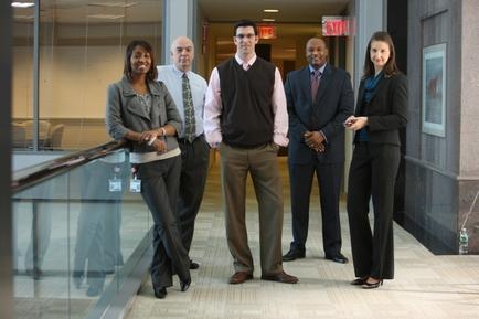 A group of five people standing in a hallway of a building. There are six people in total three men and three women all dressed in formal attire. They are standing close together and appear to be posing for a photo. The hallway has a glass railing on the left side and a large window on the right side through which we can see the interior of the building. The floor is made of marble tiles and there is a red exit sign visible in the background. The people in the photo are smiling and looking at the camera.