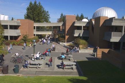 An aerial view of a large building with a large dome-shaped dome on top. The building appears to be a university or college campus as there are several other buildings visible in the background. The sky is blue and there are trees and greenery surrounding the building. In the foreground there is a courtyard with several picnic tables and benches and a group of people walking around. There are also a few bicycles parked on the left side of the courtyard. The overall atmosphere of the image is lively and bustling.