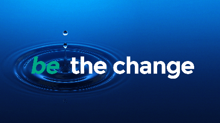 A graphic design with a blue background. In the center of the image there is a circular shape with a single drop of water falling into it. The drop is in the shape of a circle and is slightly tilted to the right. The word "be the change" is written in white capital letters in a modern sans-serif font. The overall design is simple and minimalist.