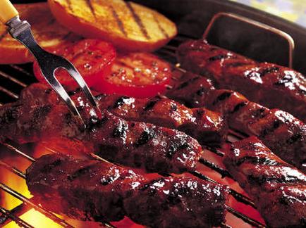 A close-up of a grill with several skewers of meat being cooked on it. The meat appears to be beef or pork with a dark brown color and a marbled texture. The skewers are arranged in a circular pattern on the grill with some of them overlapping each other. There are also some sliced tomatoes and a slice of orange on the side of the grill. A fork with a yellow handle is resting on one of the skewers. The background is blurred but it seems to be an outdoor setting with a colorful tablecloth.