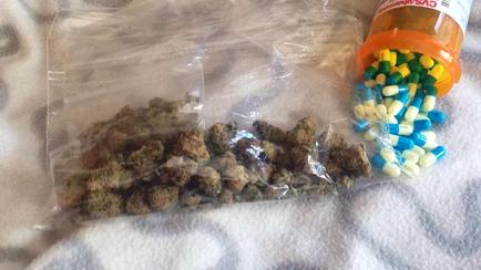 A transparent plastic bag filled with small pieces of marijuana buds. The bag is lying on a white surface with a white towel underneath. On the right side of the image there is an orange prescription bottle with a label that reads "CVS". Next to the bottle there are several colorful pills spilling out of the bottle. The pills are of different colors - blue green yellow and white.