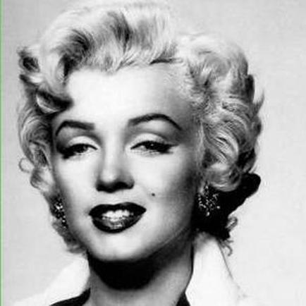 A black and white portrait of Marilyn Monroe. She is shown from the waist up looking directly at the camera with a slight smile on her lips. Her hair is styled in a voluminous curly updo with loose curls framing her face. She has a pair of large dangling earrings on her ears. The background is plain and blurred putting the focus on Marilyn's face.