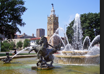 A large fountain in the center of a park. The fountain is made of bronze and is in the shape of a horse and rider. The horse is galloping forward with its mane and tail flowing in the wind. The rider is holding onto the horse's reins and appears to be in control of the water. In the background there is a tall clock tower with a clock on top. The clock tower is surrounded by trees and there are several other buildings visible in the distance. The sky is blue and the overall atmosphere of the image is peaceful and serene.