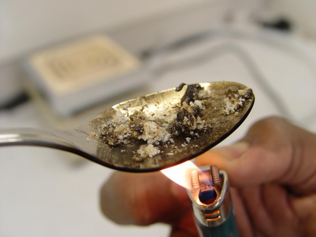 A silver spoon with a small amount of white powder on it likely an opioid or another drug. A person is holding a lit lighter under the spoon and the powder seems to be darkening in color due to the heat. The background is blurred but it appears to be light in color and possible a kitchen or laboratory setting.