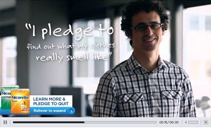 A screenshot of a video clip from a website. It shows a young man with curly hair and glasses wearing a plaid shirt standing in an office with a smile on his face. On the left side of the image there is a text that reads "I pledge to find out what my clothes really smell like" and on the right side there are two logos - one for Nicorette and the other for Rollover to expand. The background is blurred but it appears to be an indoor space with large windows.