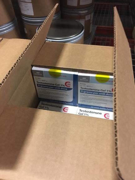 A cardboard box with two boxes inside. The box on the left is labeled "Testosterone Gel 1%" and has a blue and white label with the brand name and product name written on it. The label also has a yellow circle with a red line through it indicating that the product is a testosterone gel. The right box has a label that reads "Gel 1%". The box is open revealing the contents inside. In the background there are several other boxes and containers stacked on top of each other. The boxes appear to be in a warehouse or storage area.