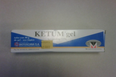 A photograph of a white and blue box of Ketum gel. The box is rectangular in shape and has a blue and white label on the front. The label has the brand name "KETUM gel" written in bold black letters at the top followed by the product information "tubo com 30g de gel - uno topico" in smaller black letters. Below that there is a description of the product which reads "Biotoscana S.A.". On the right side of the label there is a logo with a large M against a globe. The photograph is blurry. 