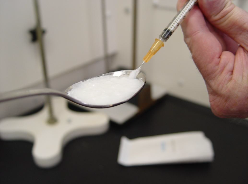 A hand holding a syringe and a spoon full of a white substance likely an opioid or another drug. The syringe is being used to draw up the substance. The background is blurred but it appears to be a laboratory setting.