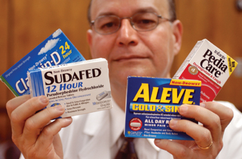 A middle-aged man wearing a white lab coat and glasses holding four boxes of over-the-counter medications in his hands. The boxes contain (from left to right) : Claritin-D Sudafed 12 Hour Aleve Cold & Sinus and Pedia Care. The background is blurry but appears to be a light brown (wood paneled?) indoor wall.