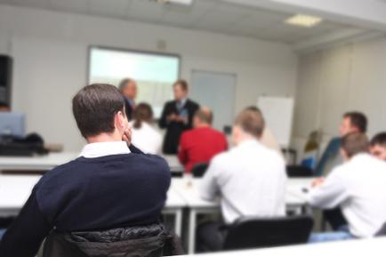 A classroom with a group of people sitting at desks and a man standing in front of a projector screen. The man is wearing a blue sweater and is sitting on a chair with his back to the camera looking towards the projector screen with a serious expression on his face. He appears to be giving a presentation or presentation to the group. There are other people in the room some of whom are sitting at the desks and some are standing. The room has white walls and a whiteboard on the right side of the image.