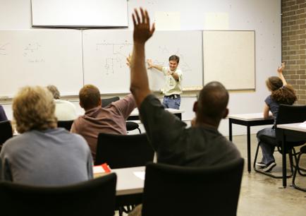 A classroom with a group of students sitting at desks and a teacher standing in front of them. The teacher is giving a presentation to the students who are raising their hands in the air. The students are attentively listening to the teacher and appear to be engaged in the presentation. The room has a brick wall in the background and a whiteboard on the wall. There are also some drawings and diagrams on the whiteboard.