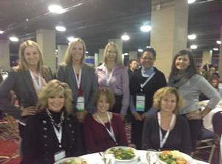 A group of nine women posing for a photo at an event. They are all dressed in formal attire and are standing around a table with plates of food in front of them. The women are smiling and appear to be happy and relaxed. The table is covered with a white tablecloth and there are other tables and chairs in the background. The event appears to be taking place in a large room with high ceilings and large windows.