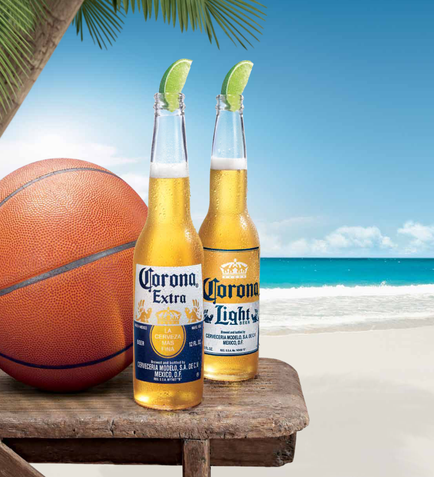 Two bottles of Corona Extra beer on a wooden table on a beach. The bottles are filled with the golden-colored beer and have a lime wedge on top. The table is surrounded by palm trees and the ocean can be seen in the background. A basketball is resting on the table next to the bottles. The sky is blue and the weather appears to be sunny and warm.