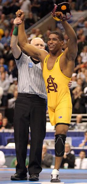 A wrestler in a yellow singlet with the letter "A" on it holding up his arms in triumph. He is standing on a wrestling mat with a referee in a black and white striped shirt standing next to him. The wrestler is smiling widely and appears to be celebrating a victory. The background shows a large crowd of spectators in the stands. The image appears to have been taken at a wrestling match.