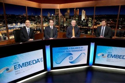Four men sitting at a desk in front of a TV studio set with a view of a city skyline in the background. The desk is set up with three large screens displaying the logo of New embed a company that provides chronic pain management services. The men are all wearing suits and appear to be in a professional setting. They are all smiling and looking at the camera. The studio has a modern and professional design with a large window that offers a panoramic view of the city skyline.