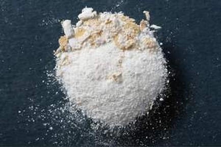 A pile of white powder on a black surface. The powder appears to be finely ground and has a rough texture. It is scattered around the pile with some pieces of the powder spilling out onto the surface. There are also some small pieces of wood shavings scattered around adding texture and depth to the image. The background is a solid black color making the powder stand out even more.