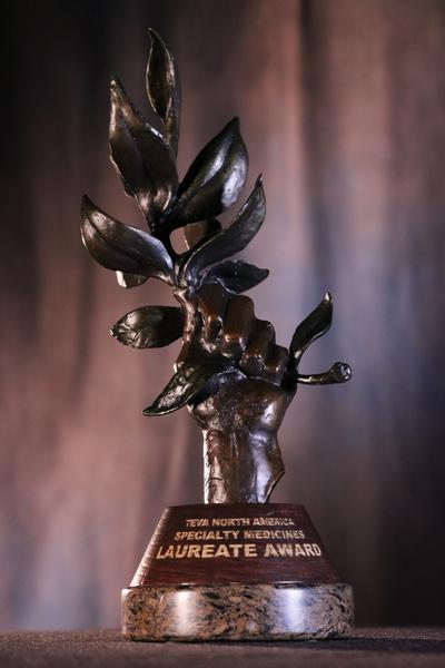 A close-up of a trophy or award. The trophy is made of bronze and has a round base with the words "Laureate Award" inscribed on it. On top of the base there is a sculpture of a hand holding a bunch of leaves. The leaves are intricately detailed and appear to be made of metal. The background is blurred making the trophy the focal point of the image.